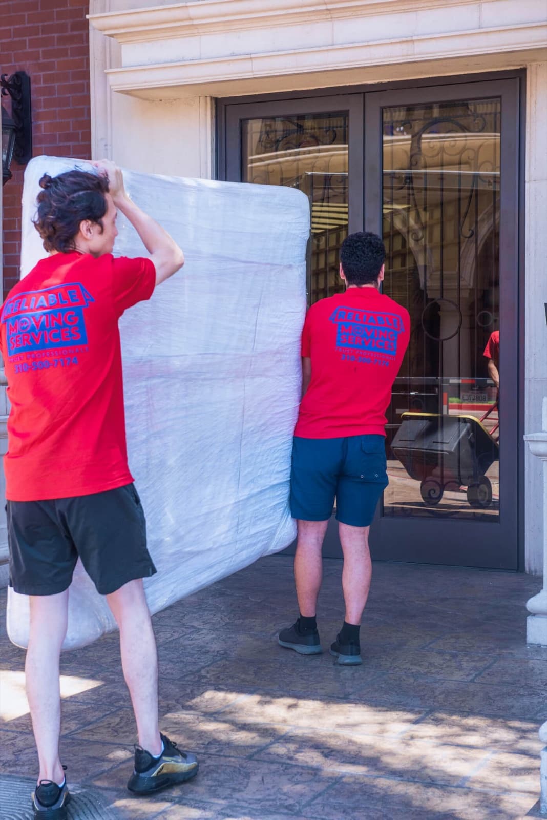 Two movers carrying a plastic-wrapped mattress into the building